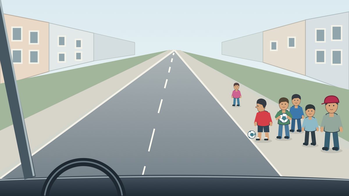 Driver view along a two-way street: six children stand on the right pavement, with one football held by the child in green and another beside the kerb near the child in red.